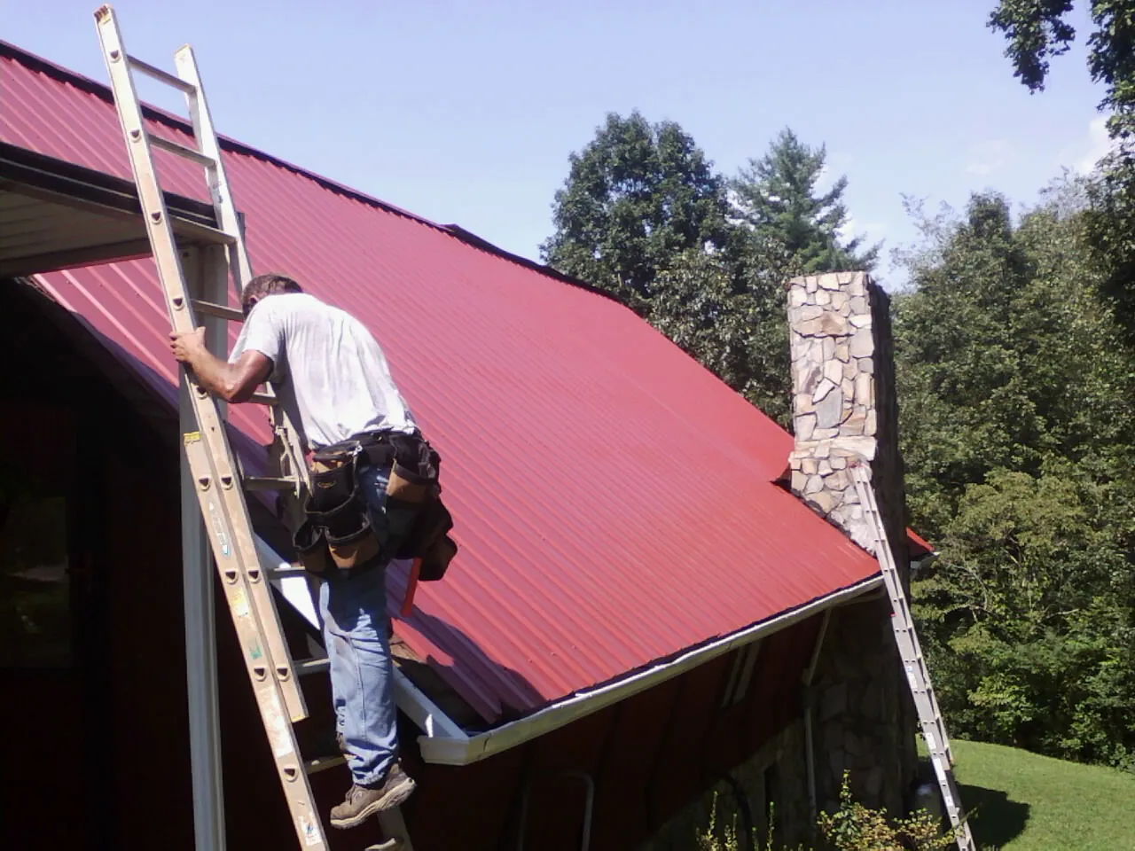 Roofer on a ladder beside a red metal roof and stone chimney — Soffit & Fascia Repair by Roofing Brothers in Birch River